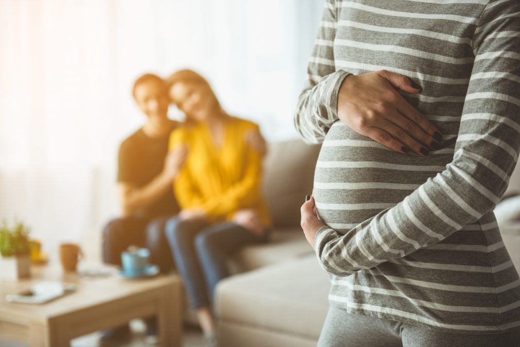 Focus on pregnant woman touching her belly. Happy married couple are sitting on sofa and embracing on background. Surrogacy concept; blog: 5 Things You Need to Know About Using a Gestational Carrier