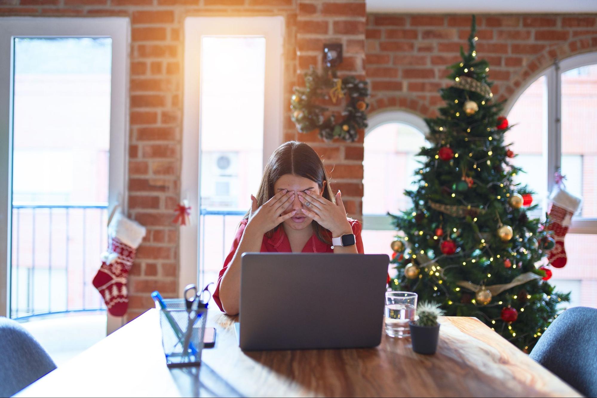 A stressed woman facing her laptop with a Christmas tree in the background to represent managing stress this holiday season