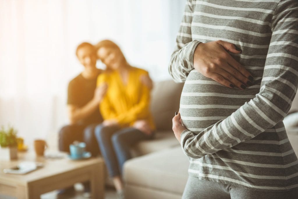 Focus on pregnant woman touching her belly. Happy married couple are sitting on sofa and embracing on background. Surrogacy concept; blog: 5 Things You Need to Know About Using a Gestational Carrier